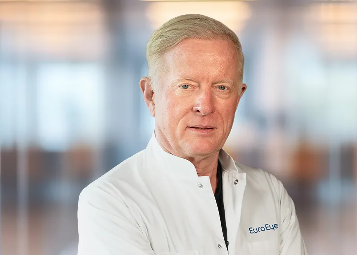 Porträt von Dr. med. Jørn S. Jørgensen s in einem weißen Arztkittel mit dem Logo ‚EuroEye‘ auf der Brust. Er blickt ruhig und freundlich in die Kamera, steht leicht seitlich, Arme locker verschränkt. Der Hintergrund ist hell und unscharf, erinnert an eine moderne Klinik oder Praxisumgebung.