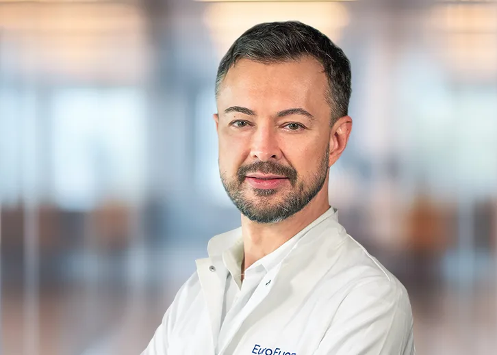 Porträt eines männlichen Arztes in halber Länge, Blick nach vorn. Er trägt einen weißen Arztkittel mit dem Schriftzug ‚EuroEyes‘. Sein Gesicht ist freund­lich, gepflegt, mit kurzem dunklem Haar und dezentem Bart. Der Hintergrund ist unscharf, mit hellen, weichen Farbtönen.