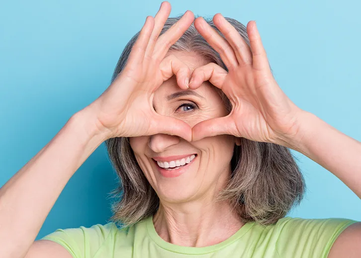 Nach der Operation Eine ältere Dame strahlt in die Kamera und formt mit ihren Fingern ein Herz. Sie hält das Herz über ihr Auge.